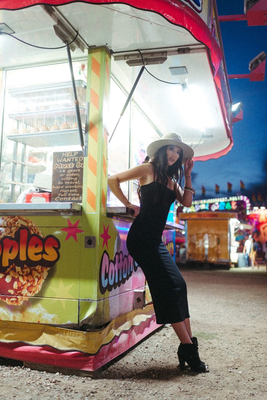 Trendy, stylish young woman in black dress and hat leans weight against carnival fair food truck 
