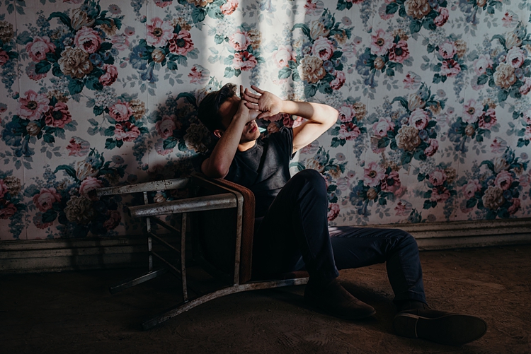 Young man sits against wallpapered, floral wall on over-turned chair blocking sun from his eyes